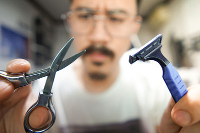 Man wearing eyeglasses holding scissors and razor at home