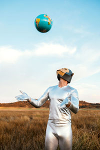 Man in geometric monkey mask and silver latex suit tossing earth painted ball in yellow grassy field in sunny day