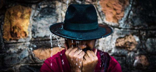 Close-up of man wearing hat against wall during rainfall