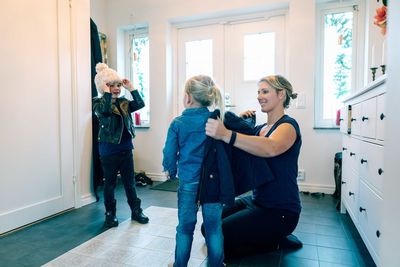 Smiling mother helping daughter with jacket while kneeling at home