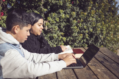 Two young latino students, outdoors, using laptops, sitting at a park