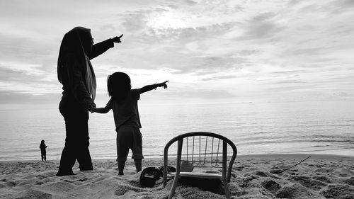 Surface level of mother and daughter gesturing while standing on sand at beach against sky