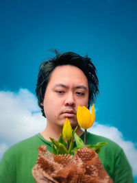 Portrait of young man holding yellow tulips against cloudy blue sky.