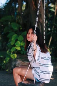 Portrait of young woman sitting on swing against tree