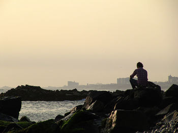 Rear view of man sitting on rock formation by sea