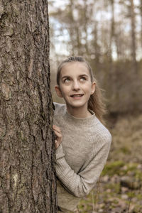 Smiling teenage girl looking up while standing behind tree in forest