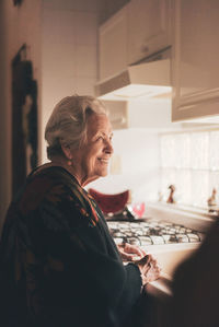 Side view of happy positive senior female with gray hair wearing warm clothes standing at sink in kitchen