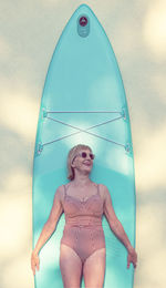 Woman in a swimsuit and sunglasses stands in front of a stand-up paddleboard