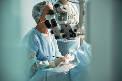 Focused adult female doctor in sterile mask and ornamental medical cap looking through surgical microscope against crop coworker in hospital