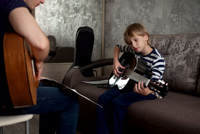 Young male musician teaches little boy student how to play the acoustic guitar and play by notes.