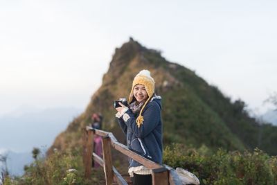Portrait of smiling woman holding camera against mountain