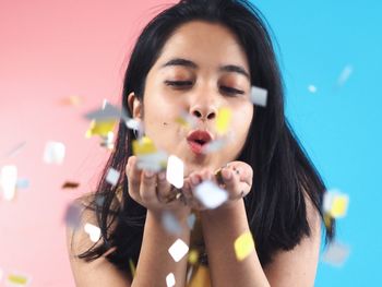 Young woman blowing confetti against pink and blue background