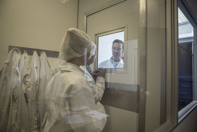 Two lab technicians in sterile protective clothing looking at colleague behind glass pane