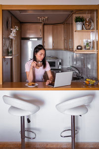 Focused asian female with hot drink surfing netbook while sitting at counter with food during breakfast in modern kitchen at home