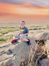 Portrait of boy sitting on sand at beach against sky during sunset