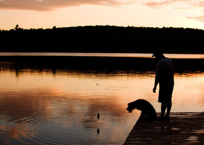 Silhouette of man with dog on water at sunset