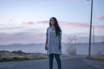 Young woman looking away while standing on road against sky during sunset