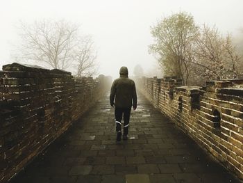 Rear view of man walking on footpath by brick wall against foggy sky
