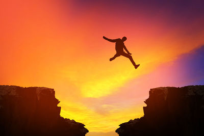 Low angle view of silhouette man jumping on cliff against sky during sunset