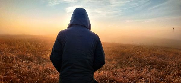 Rear view of man wearing hood standing on field against sky during sunset