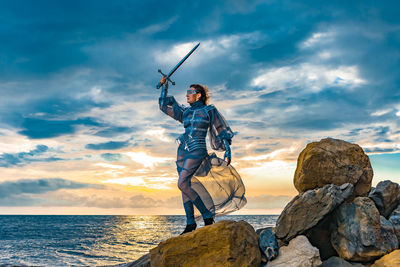 Full-length portrait of a woman medieval knight standing on a rock against a dramatic sky and a sea 