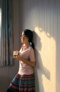 Thoughtful woman drinking coffee while standing by wall at home