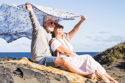 Happy senior couple with scarf sitting at beach against sky