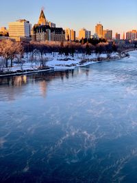 Frozen river by buildings against clear sky during sunset