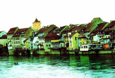 Houses by river against buildings in city against clear sky