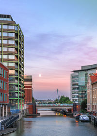 Buildings by river against sky in city at sunset