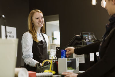 Smiling female owner looking at customer while standing in checkout counter of store