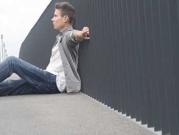 Side view of young man sitting by railing on footpath