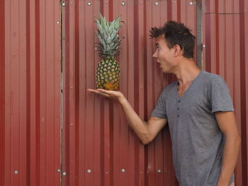 Surprised young man looking at pineapple on palm by metal sheet