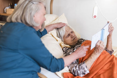 Woman taking care of old woman lying in bed reading book