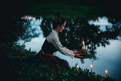 Side view of young woman holding candle sitting by lake 