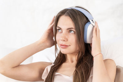 Caucasian young woman with headphones listens to music while sitting in bed in beige silk pajamas