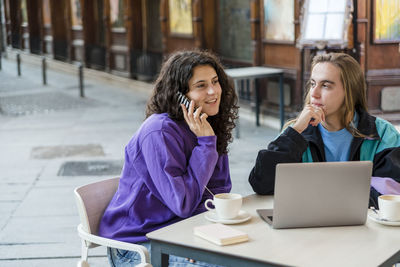 Two people using laptop and mobile phone while sitting outdoors at a coffee shop