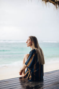 Young woman sitting on shore at beach against sky