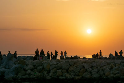 People on rocks by sea against sky during sunset