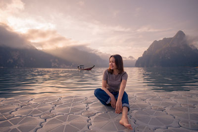 Woman sitting on shore against sky during sunset