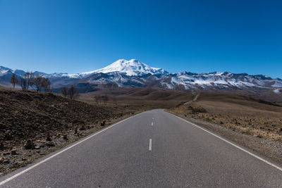 Road amidst snowcapped mountains against clear blue sky