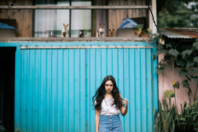 Sad pensive teen female in casual clothes touching hair while standing against countryside house with blue shutters and little dogs