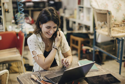 Smiling female upholstery worker using laptop while talking on phone at workbench in workshop