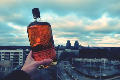 Cropped hand holding alcohol bottle in city against cloudy sky