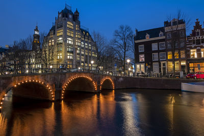 Bridge over river against illuminated buildings at dusk