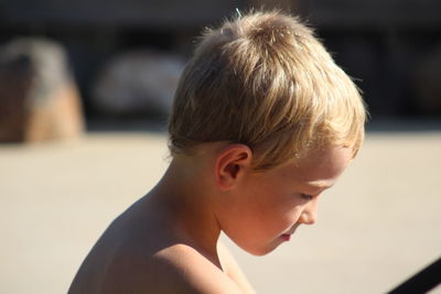 Side view of shirtless cute boy looking down while sitting at beach