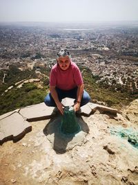 High angle portrait of man crouching on mountain against cityscape