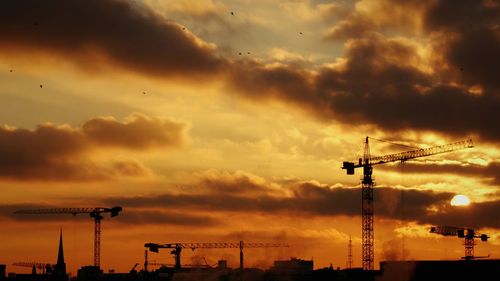 Silhouette cranes against cloudy sky during sunset