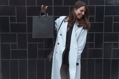 Smiling woman holding bag against wall outdoors