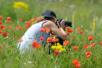 Side view of woman photographing with digital camera by flowers on field